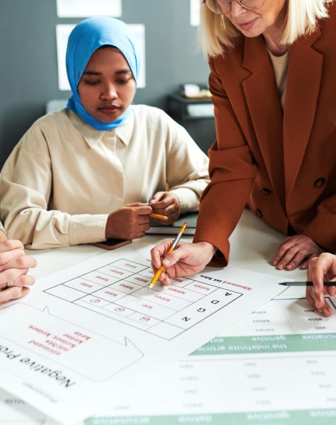 Mature blond teacher bending over document with table of pronouns while explaining new subject to group of intercultural students